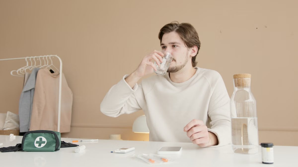 Man in white long sleeves drinking water from glass at home with medical supplies.